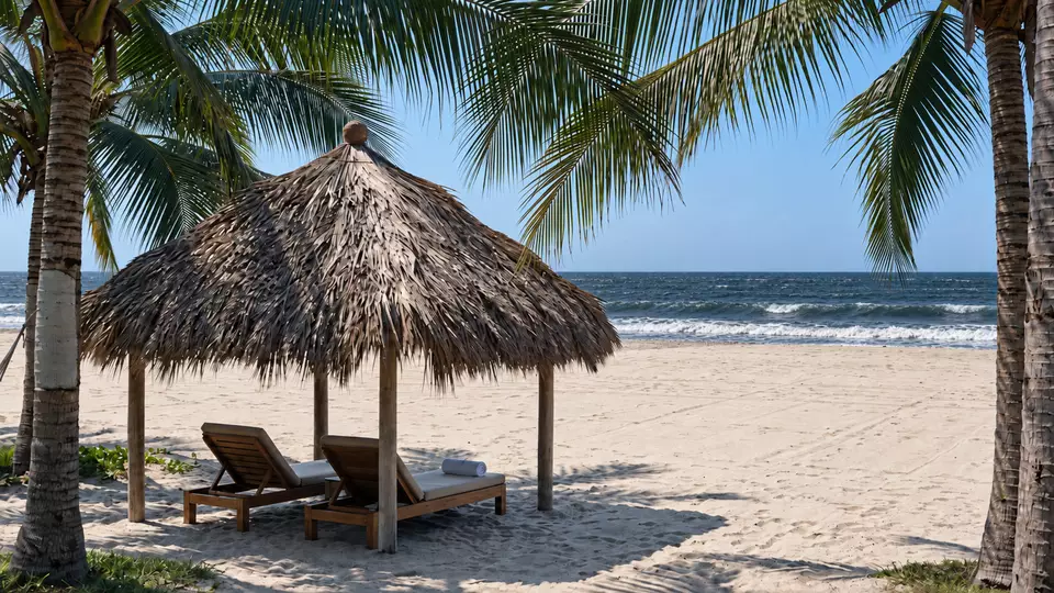 Dos tumbonas de madera bajo una palapa de palma, en arena blanca entre palmeras, frente al mar.