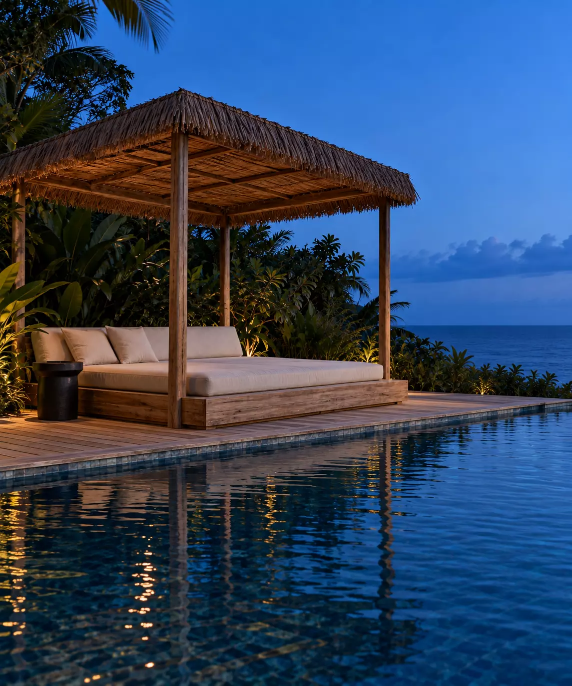 Cama exterior bajo techo de palma junto a una piscina, con el mar al fondo al anochecer.
