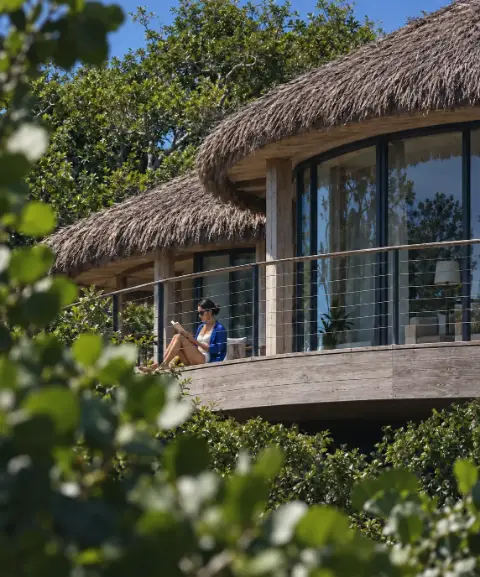 Une personne lit sur la terrasse arrondie d'un bungalow au toit de palme, entouré d'arbres.