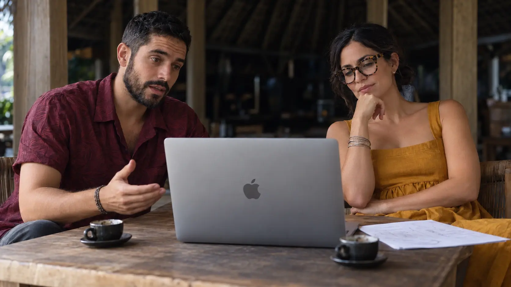 Un homme et une femme consultent un ordinateur portable autour d'un café, à une table en bois rustique sous un toit de palme.