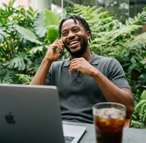 Un homme sourit au téléphone devant un ordinateur portable, entouré de plantes tropicales.