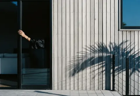 Un bras paré d'un bracelet doré ouvre une porte coulissante sombre à côté d'une façade en bois clair marquée par l'ombre d'une palme.