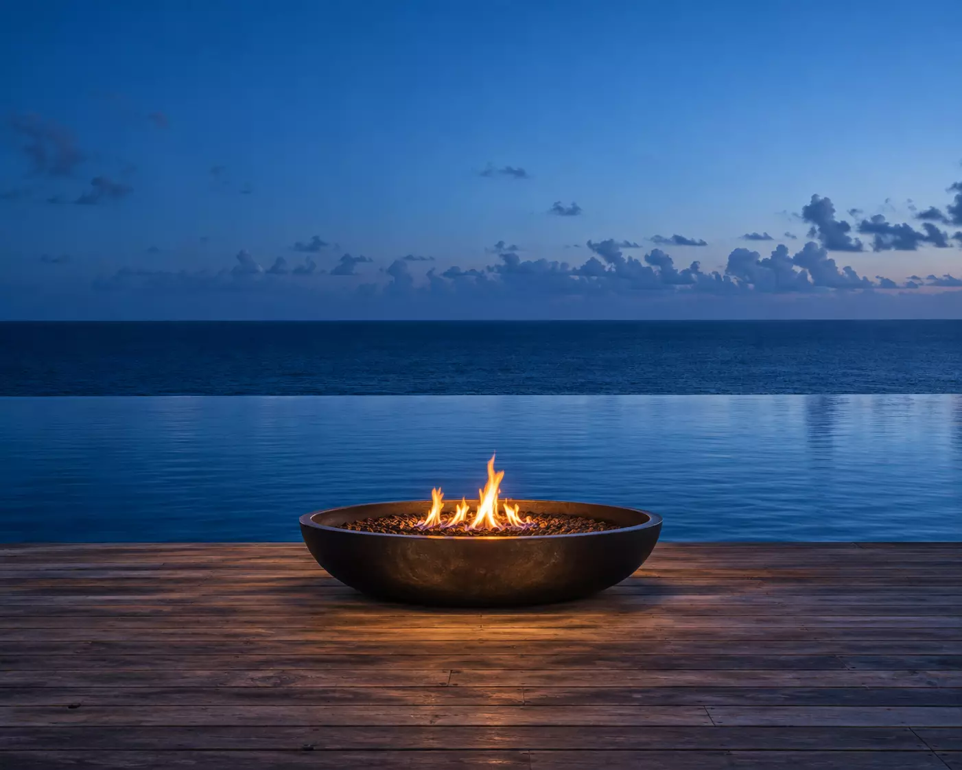 Un fuego encendido en un cuenco sobre una terraza de madera, frente al mar al anochecer.