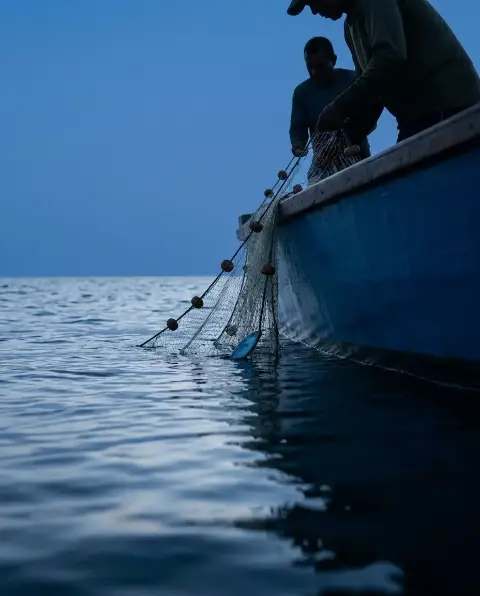 Deux pêcheurs relèvent un filet depuis une barque bleue sur une mer calme, au crépuscule.