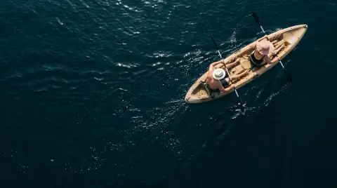 Vista aérea de dos personas con sombrero remando en un kayak sobre el mar azul oscuro.
