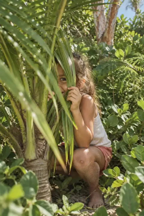 Una niña se asoma entre hojas de palma.