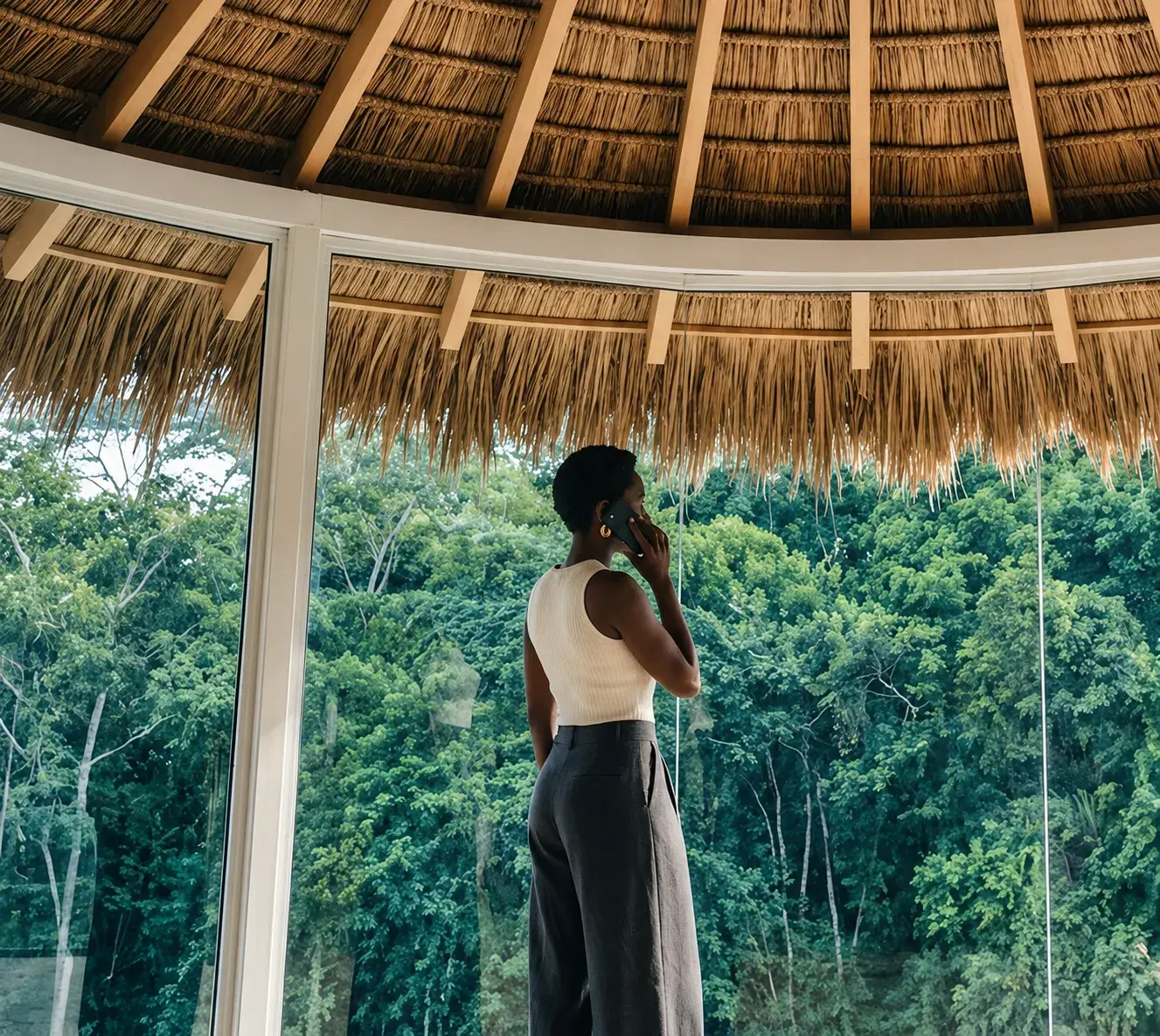 Une femme, de dos, parle au téléphone dans une pièce ronde au toit de palme, face aux baies vitrées courbes et à la végétation.