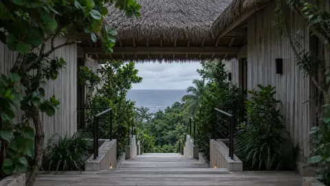 Pasarela de madera entre dos volúmenes de techo de palma, con escaleras que bajan entre la vegetación hacia el mar.
