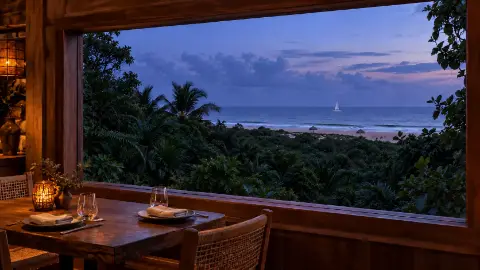 Mesa servida junto a una ventana amplia que mira hacia la vegetación y el mar, al anochecer.