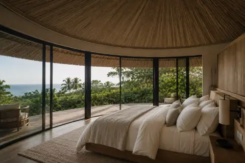 Habitación con cama amplia bajo techo de palma, rodeada de ventanales hacia la vegetación y el mar.