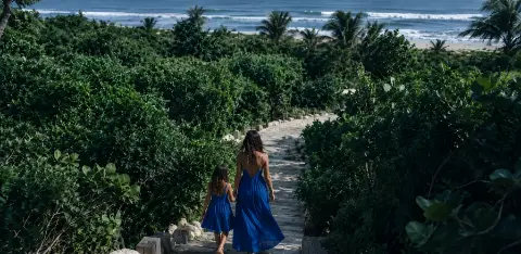 Una mujer y una niña bajan por un camino de piedra entre la vegetación hacia la playa.