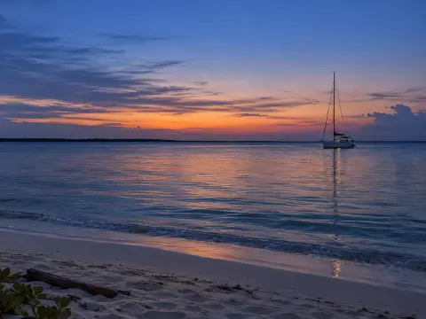 Un velero anclado frente a una orilla de arena, bajo un cielo de atardecer.