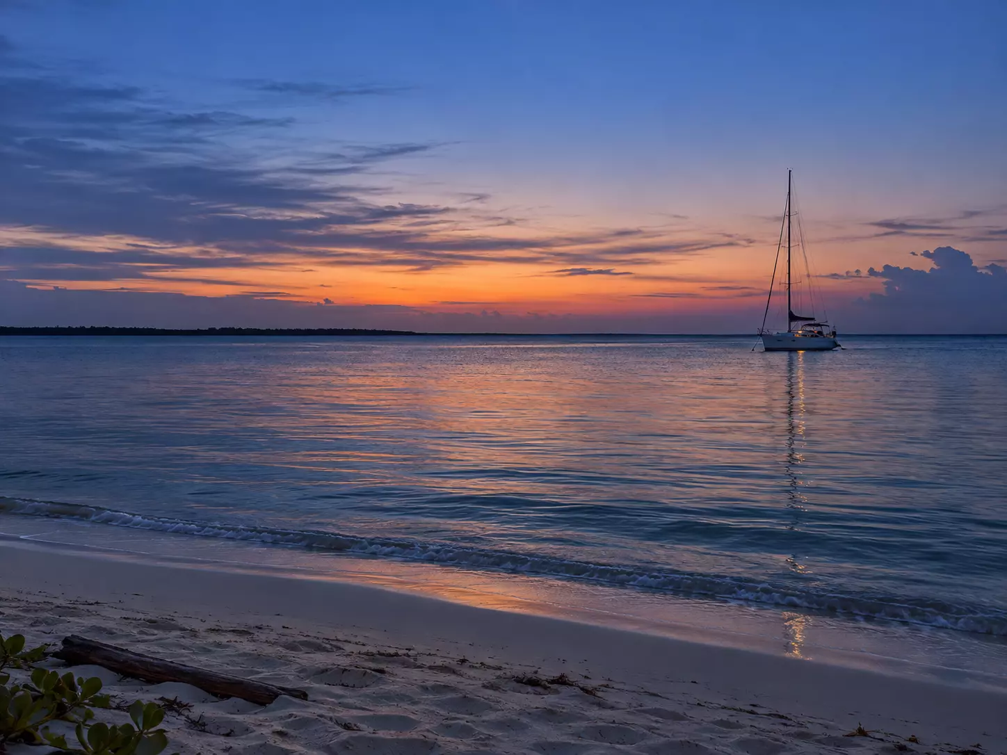 Un voilier au mouillage face à un rivage de sable, sous un ciel de coucher de soleil.