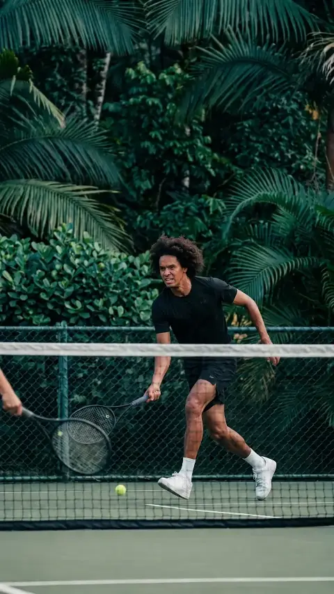 Un jugador corre hacia la red en una cancha de tenis rodeada de palmas y vegetación.