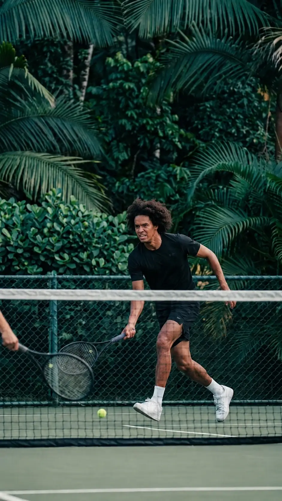 Un jugador corre hacia la red en una cancha de tenis rodeada de palmas y vegetación.